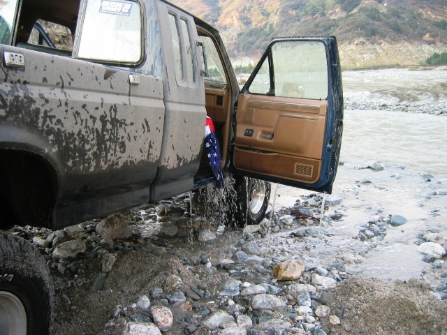 Mud-covered SUV with open door parked in a rocky, flowing riverbed