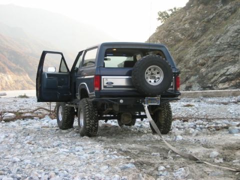 1985 Ford Bronco - This lifted Ford Bronco is parked in a rocky, outdoor area with its doors open and a spare tire mounted on the rear.
