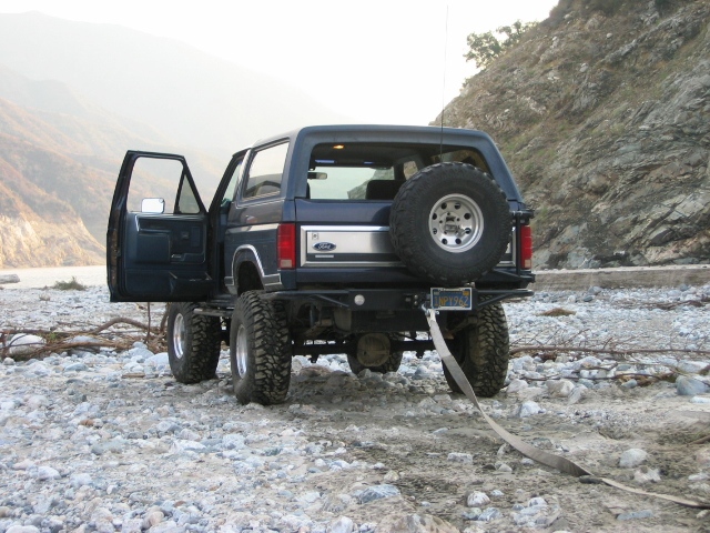 Dark blue Ford Bronco rear 3/4 view, lifted with spare tire, parked on rocky riverbed