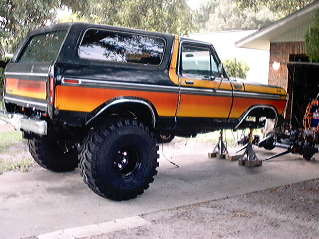 Black and orange Ford Bronco on jack stands, undergoing repairs. Large off-road tires are visible