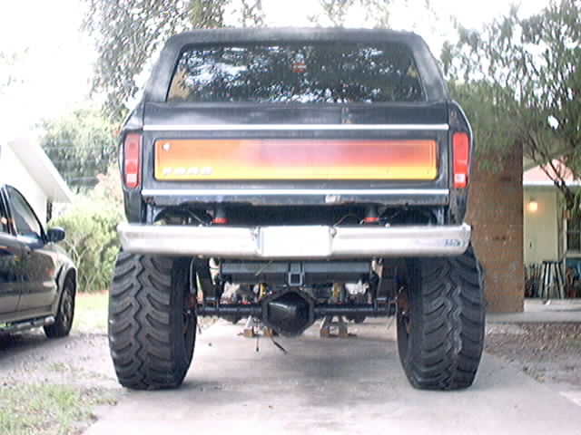 Rear view of a lifted black Ford Bronco with large tires and a red-yellow gradient taillight panel. Another car is parked to the left