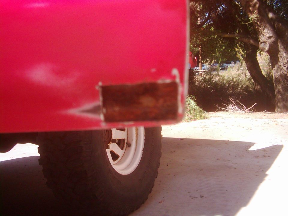 Red vehicle body panel with visible rust damage on the lower rear corner. A white wheel and tire are partially visible
