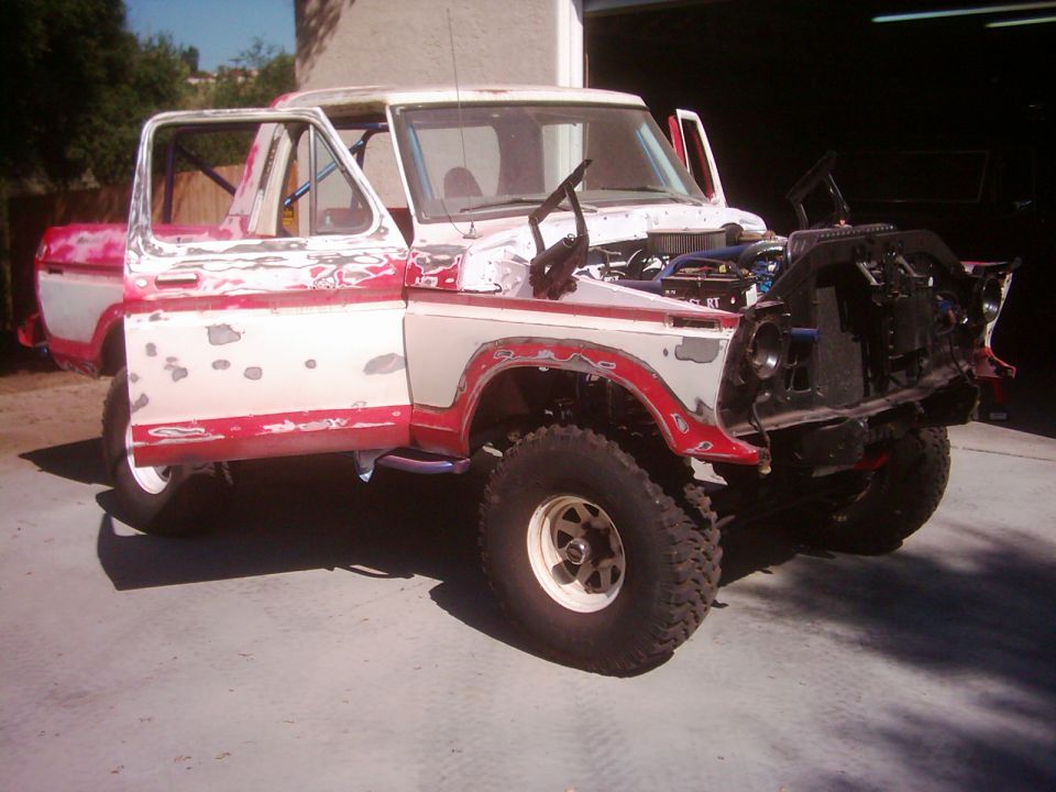 1978 Ford Bronco with doors open, hood up, and front end disassembled. White and red paint is peeling