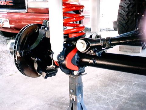 1979 Ford Bronco - Detail of vehicle suspension components, with a partial view of a red GMC truck in the background.