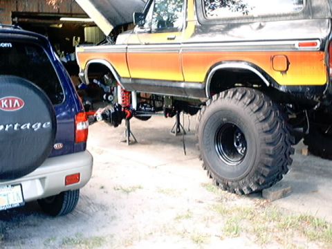 1979 Ford Bronco - A lifted 1978 Ford Bronco is undergoing repairs, with its front end elevated and axle removed, parked beside a Kia Sportage.