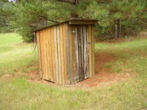 1995 Ford Bronco - A rustic wooden outhouse is situated in a natural outdoor setting surrounded by grass and trees.