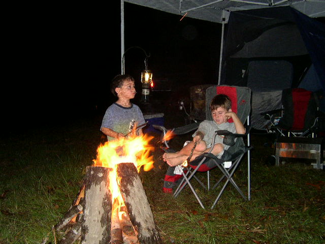 Two boys sit by a campfire at night, one roasting something over the flames, the other in a camping chair