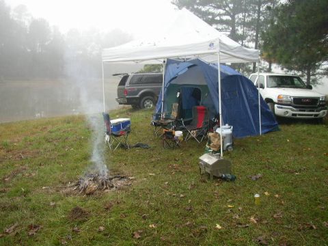 1995 Ford Bronco - A campsite is set up near a body of water with a tent, chairs, and a campfire. Two vehicles are parked nearby.