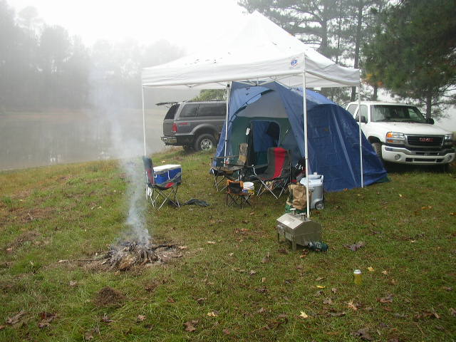 Camp site by a lake with a tent, campfire, and two trucks. One is a dark SUV, the other a white GMC pickup
