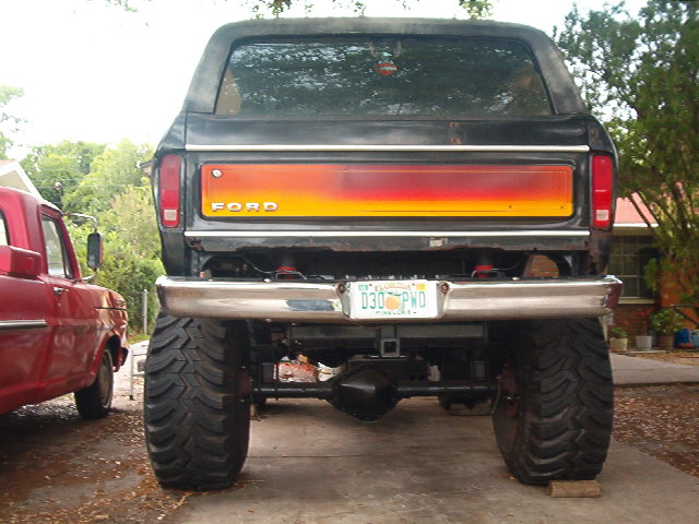1979 Ford Bronco rear view, lifted with large tires and custom tailgate paint. Red truck parked to the left