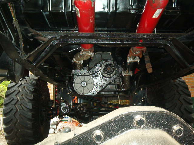 Underbody view of a vehicle with large tires, drivetrain, and red suspension components