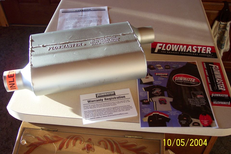 Silver Flowmaster muffler with INLET label beside warranty and apparel paperwork on a table