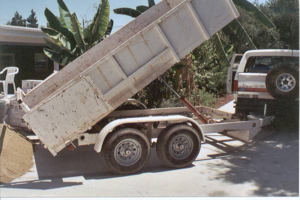 White tandem-axle dump trailer with bed raised dumping sand behind a white SUV