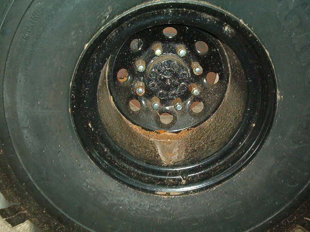 Close-up of a black wheel with a large tire. The wheel has multiple lug nuts and circular cutouts. Some rust is visible on the wheel's inner rim