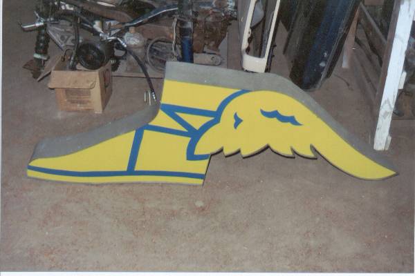 Yellow winged-foot logo sign cutout lying on a garage floor with parts and a box behind it