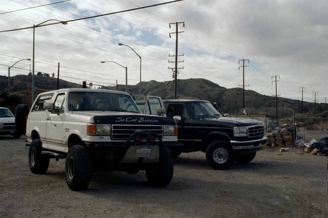 Lifted white Ford Bronco with 'So Cal Broncos' hood lettering parked beside a dark Ford pickup in a dirt lot