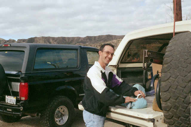 Man at open tailgate of white SUV with rear-mounted spare tire, dark blue SUV parked beside it in desert setting