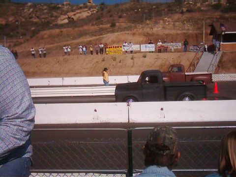 1978 Ford Bronco - Two pickup trucks race down a drag strip with a crowd watching from the hillside.