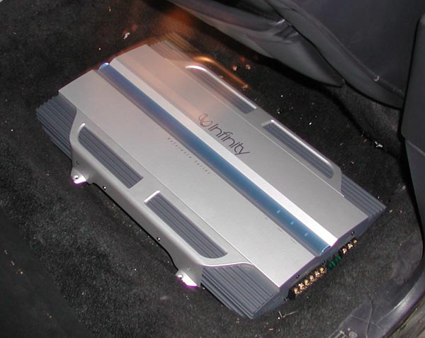 Silver Infinity car audio amplifier with gold terminals sitting on dark vehicle carpet