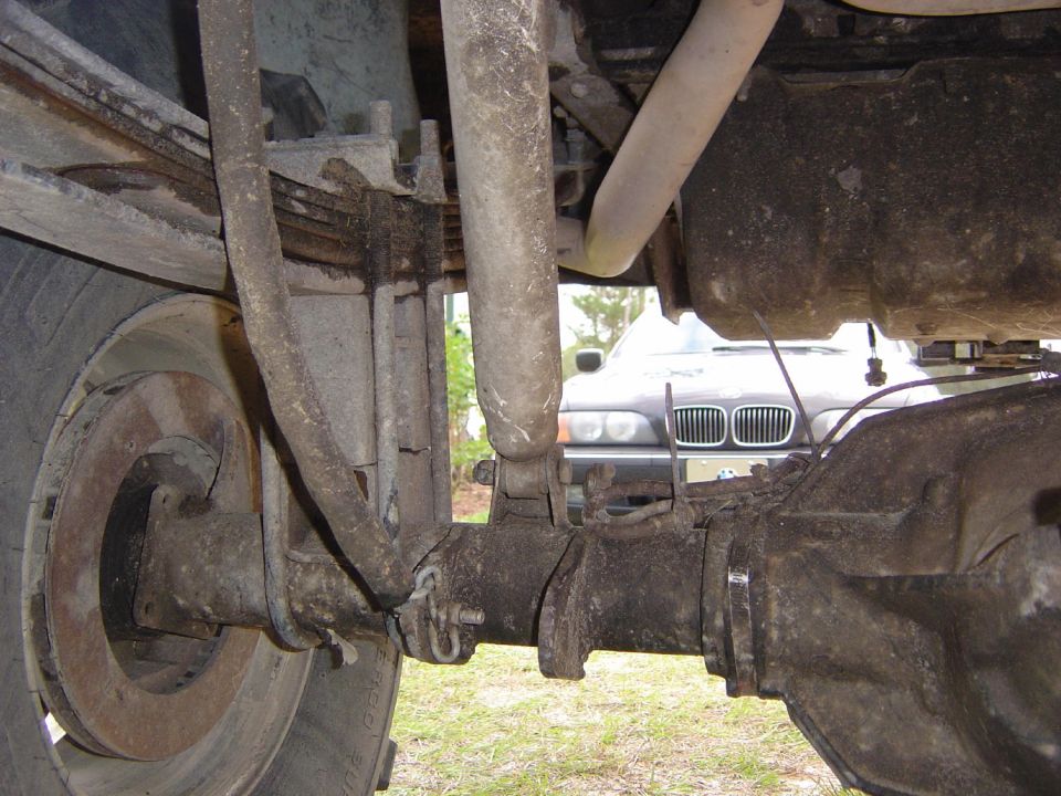 Close-up of a vehicle's rear axle, leaf springs, and shock absorber. Another car is visible in the background