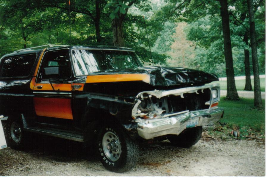 Black 1970s Ford Bronco with orange stripes and crushed front end on a gravel driveway