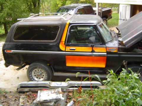 1979 Ford Bronco - This Ford Bronco has a distinctive orange stripe and a roof rack, with a pickup truck visible behind it.