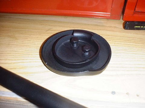 1988 Ford Bronco - A black metal component, possibly a cap or bracket, with two bolts is shown on a wooden workbench.