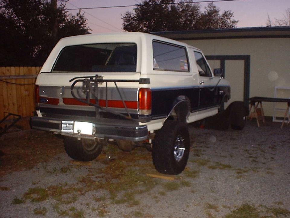 White and blue Ford Bronco with a rear spare tire carrier, lifted suspension, and chrome wheels