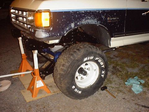 1988 Ford Bronco - The front axle of a Ford Bronco is supported by jack stands, with large tires visible.