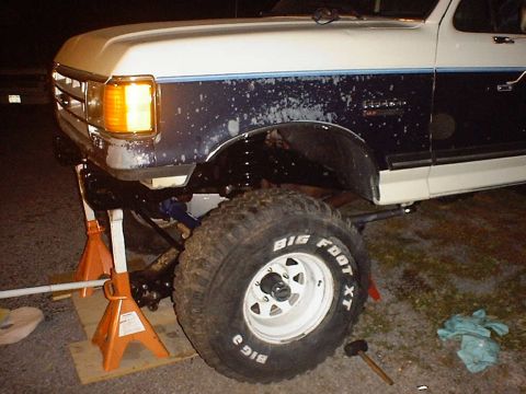 1988 Ford Bronco - The front axle and tire of a lifted truck are visible, with the vehicle supported by orange jack stands.