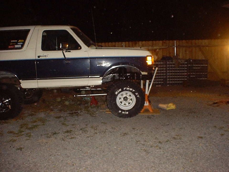 Side view of a lifted Ford Bronco with "Bigfoot XT" tires, supported by jack stands at night