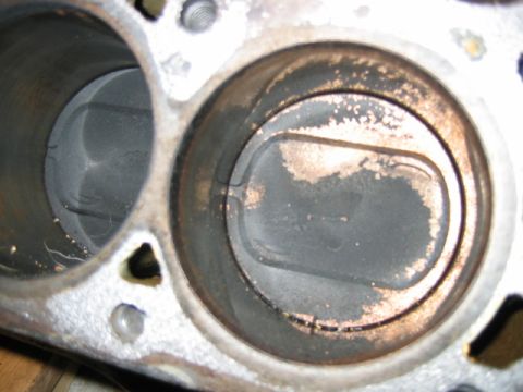 1992 Ford Bronco - Close-up of two engine cylinder bores in an aluminum block or head, with the right piston crown visible showing carbon buildup, discolorati…