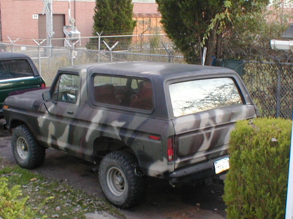 Camouflage-painted 1970s Ford Bronco parked by a chain-link fence, rear view