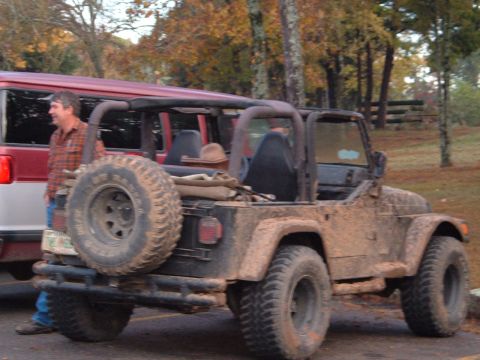 1986 Ford Bronco - A man stands next to a red SUV while a muddy Jeep Wrangler is parked beside it, showing off its off-road capabilities.
