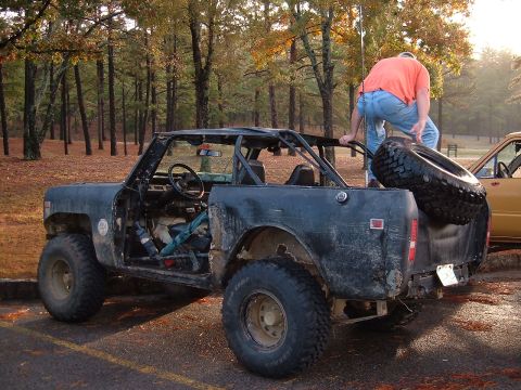 1986 Ford Bronco - A black International Scout II with a roll cage is seen with a person loading a spare tire into the bed, parked next to another vehicle.