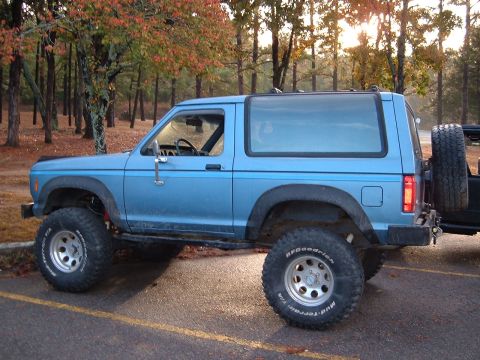 1986 Ford Bronco - A lifted blue 1986 Ford Bronco is parked outdoors with trees in the background, its off-road capabilities.