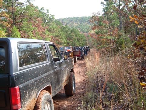 1986 Ford Bronco - Several Ford Broncos driving in a line on a dirt road through a wooded area.