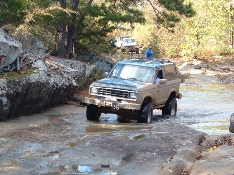 1986 Ford Bronco - A 1986 Ford Bronco navigates a rocky stream during an off-road excursion, with other vehicles following in the background.