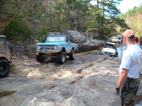 1986 Ford Bronco - Several off-road vehicles traverse a challenging rocky path with trees and boulders surrounding them.