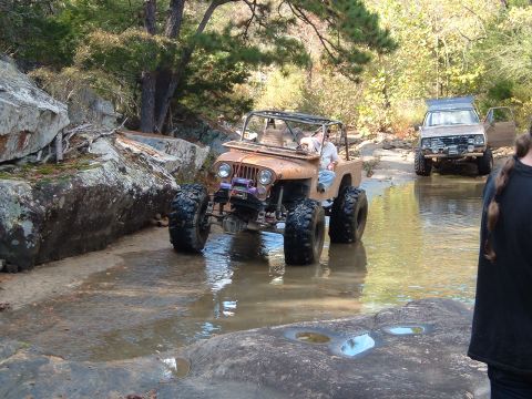 1986 Ford Bronco - Two modified off-road vehicles traverse a shallow, rocky stream surrounded by trees and boulders.