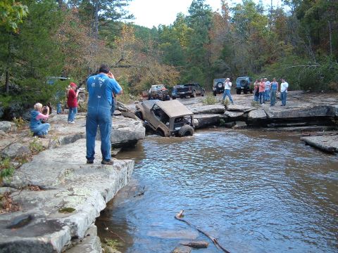 1986 Ford Bronco - Several off-road vehicles are crossing a shallow stream in a wooded area, with people observing.