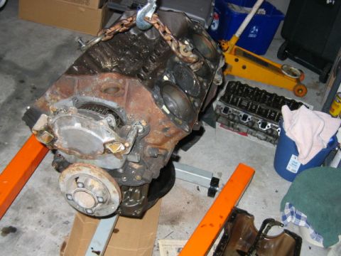 1992 Ford Bronco - A rusty engine block sits on an orange engine stand or lift arms in a garage, with a chain hoist attached to its lifting eye. In the backgr…