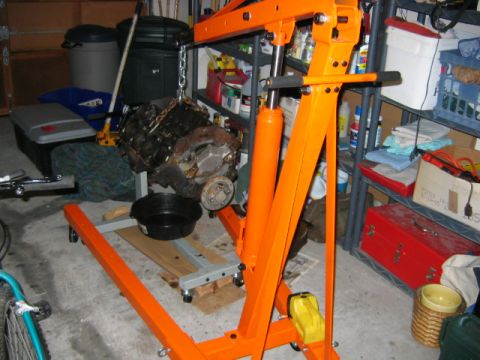1992 Ford Bronco - An engine block hangs suspended from an orange hydraulic engine hoist (cherry picker) with a chain, sitting in a cluttered garage. The grea…