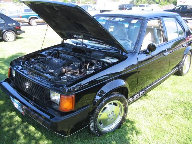 Black Shelby Dodge Omni hatchback with hood up showing engine, parked on grass at a car show