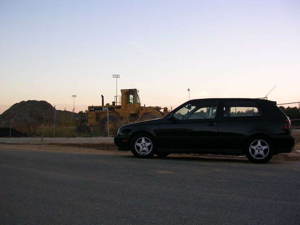 Black Volkswagen hatchback parked on asphalt with a yellow Caterpillar loader in the background