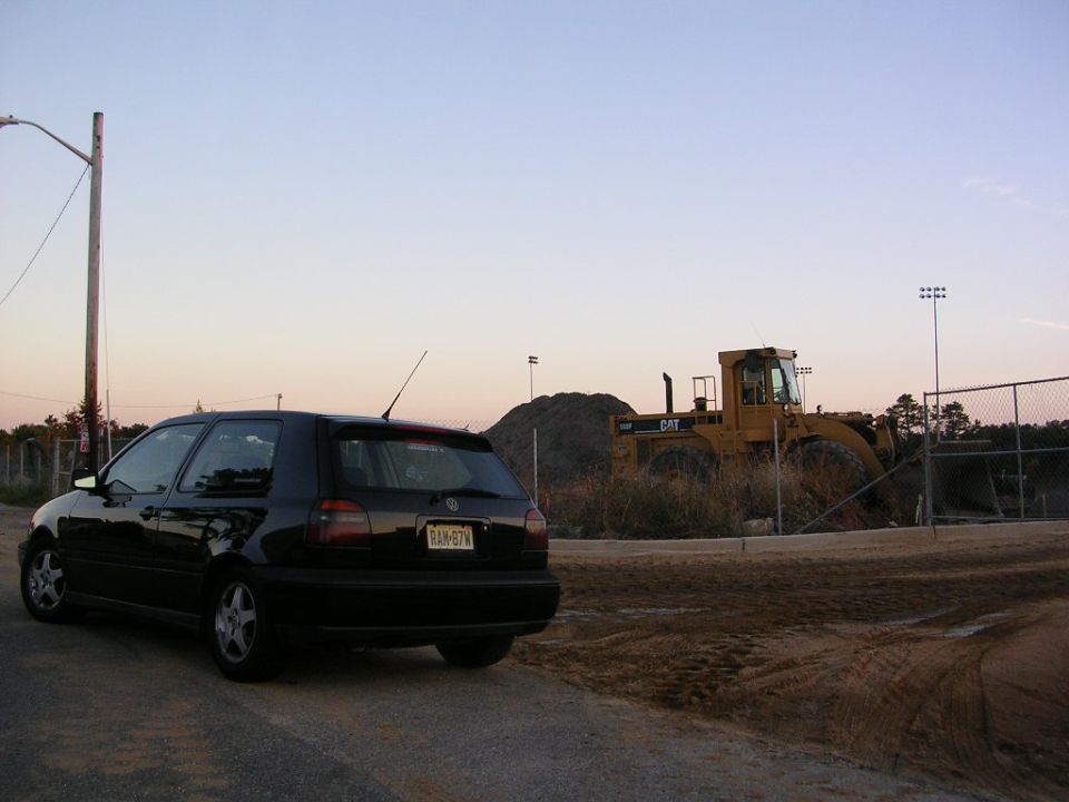 black hatchback parked on dirt road, yellow CAT front loader in background