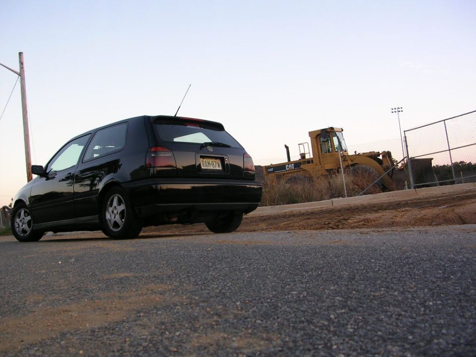 Black VW GTI hatchback rear view on pavement, CAT loader behind fence at dusk