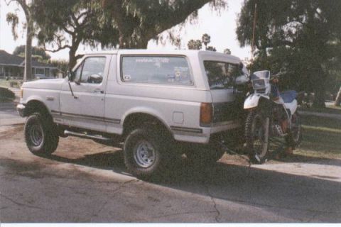 1990 Ford Bronco - A white 1990 Ford Bronco is parked on a street with a dirt bike loaded on a trailer behind it.