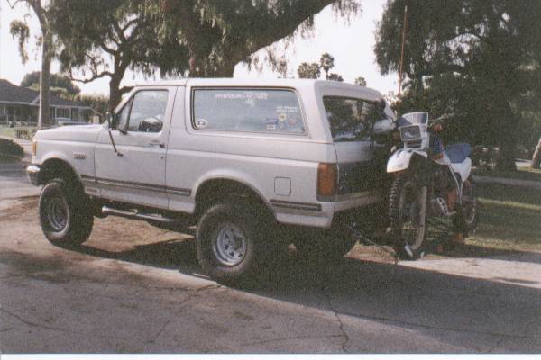 White 1990 Ford Bronco with a dirt bike on a trailer, parked on a street