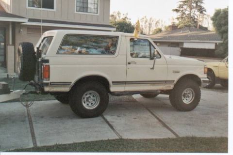 1990 Ford Bronco - A white Ford Bronco is parked on a driveway next to a yellow vehicle, with a house in the background.